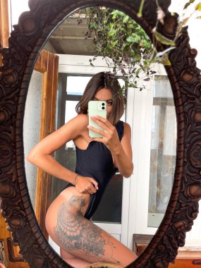 Evdeki güzel kadın cep telefonundaki fotoğrafların aynada fotoğrafını çekiyor sosyal medyada, dikey çerçevede, siyah yaz mayosu giyiyor, bacağında büyük bir dövme var, formda ve bronzlaşmış.