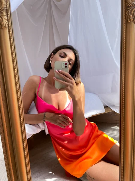 在家里穿着鲜艳的夏橙丝裙的美发女郎在手机上拍照 为社交媒体 垂直拍摄自己的照片 — 图库照片
