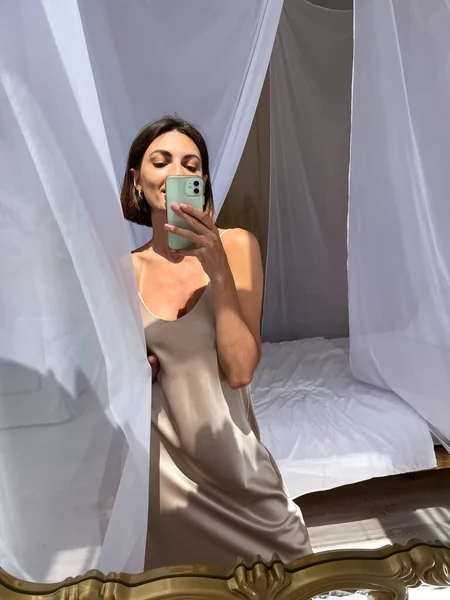 在家里穿着浪漫米色丝绸衣服的晒黑的女人在手机上拍照 在镜子里为社交媒体 垂直拍摄 — 图库照片