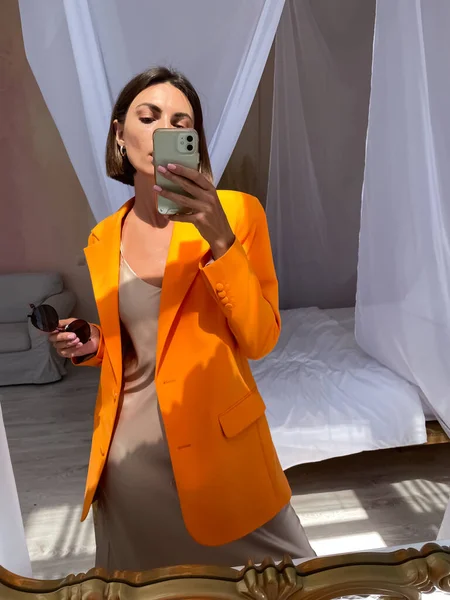 フィット日焼けした女性でロマンチックなベージュシルクドレスとオレンジブレザーで自宅取る写真自撮りで電話でミラーのためにソーシャルメディア — ストック写真