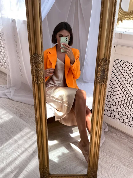 在家里穿着浪漫米黄色丝绸衣服和橙色夹克的晒黑的女人在手机上拍照 在社交媒体 垂直的镜子里拍照 — 图库照片
