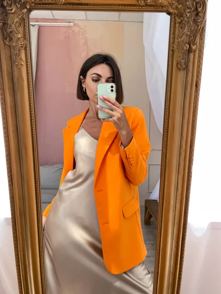 Bej rengi ipek elbiseli, turuncu ceketli, bronz tenli bir kadın sosyal medya, hikayeler, dikey dikey görüntüler için telefonda fotoğraf çekiniyor.