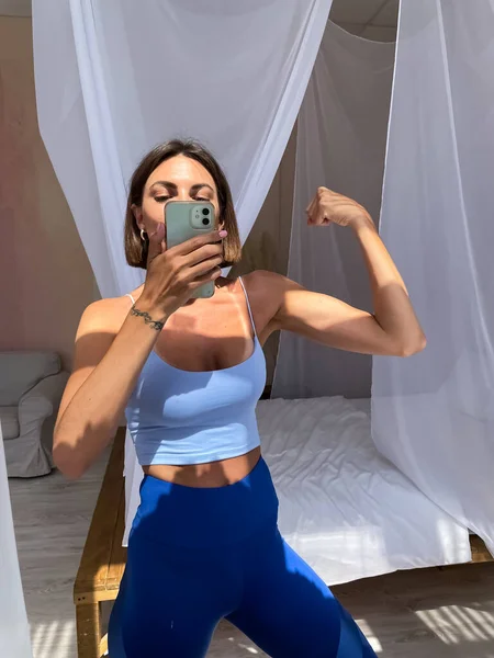 Spor kıyafetleri içinde bronzlaşmış kadın, mükemmel vücut, karın kasları motivasyonu, evde sosyal medya, hikayeler, dikey dikey dikey görüntüler için telefonda fotoğraflı selfie çek.