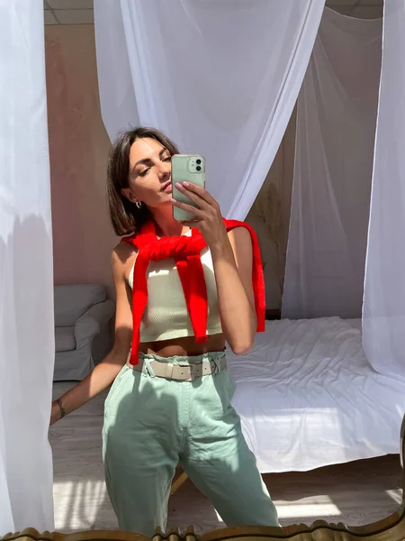 在家里穿着鲜艳的红色毛衣和薄荷绿色裤子的时尚女性在手机上拍照 这是在手机上的社交媒体上拍摄的 — 图库照片