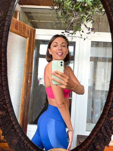 Evdeki güzel kadın cep telefonundaki aynada fotoğraf çekiniyor sosyal medyadaki haber ve paylaşımlar için, dikey çerçeve, spor kıyafetleri, forma ve ince, karın kasları motivasyonu