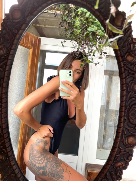 在家里 漂亮的女人穿着黑色夏装 腿上有个很大的纹身 适合晒晒黑衣服 在社交媒体上的故事和贴文都是在手机上拍照的 — 图库照片