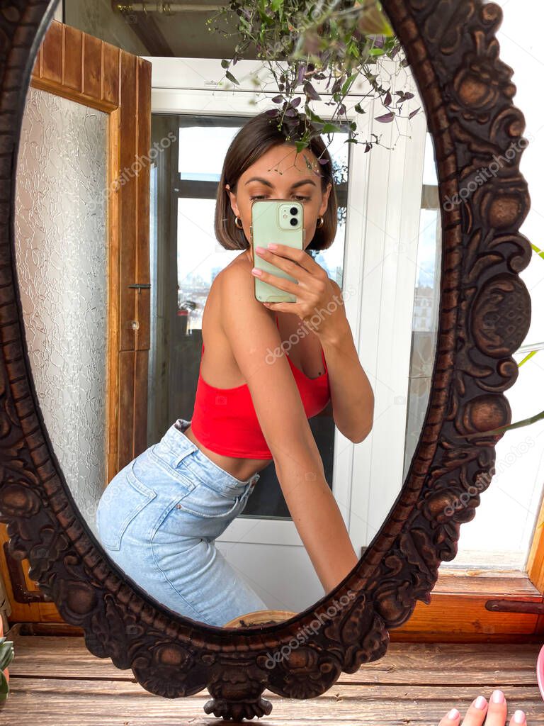 Pretty woman at home take photo selfie in mirror on mobile phone for stories and posts in social media, vertical frame, wearing jeans and red top, fit slender, good looking