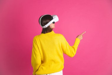 Sanal gerçeklik gözlükleriyle pembe arka planda parlak sarı süveter giyen güzel bir kadın kamerayı parmağıyla boş yere çevirmiş.
