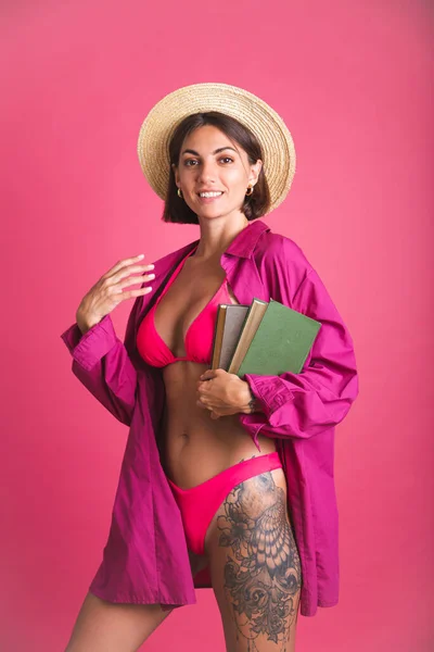 穿着比基尼和衬衫 穿着漂亮的晒黑了的运动衫的女人拿着书去海滩读粉色背景的书 — 图库照片