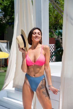 Bahçede bikinili, havuz başında hasır şapkalı güzel, bronzlaşmış bir kadın.