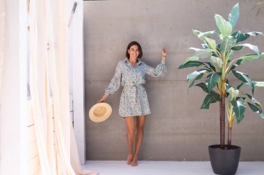 Yazlık elbise ve hasır şapka giymiş güzel bir kadın. Arka planda gri bir duvar. Neşeli, neşeli, mutlu, neşeli, neşeli, mutlu bir kadın.