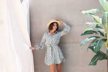 Yazlık elbise ve hasır şapka giymiş güzel bir kadın. Arka planda gri bir duvar. Neşeli, neşeli, mutlu, neşeli, neşeli, mutlu bir kadın.