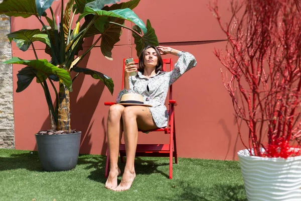 Yazlık elbiseli güzel bir kadın güneşli bir günde arka bahçede sandalyede oturuyor. Cep telefonuyla.