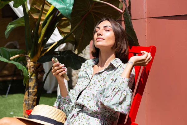 Yazlık elbiseli güzel bir kadın güneşli bir günde arka bahçede sandalyede oturuyor. Cep telefonuyla.