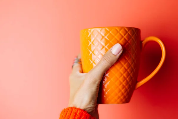 Woman\'s hand in red sweater holds orange  mug on red background