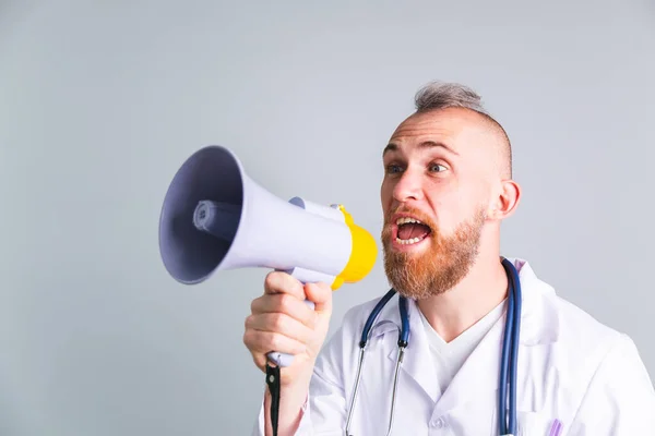 Gri arka planda yakışıklı erkek doktor megafonla bağırıyor dikkat istiyor, herkesi aşı olmaya çağırıyor.