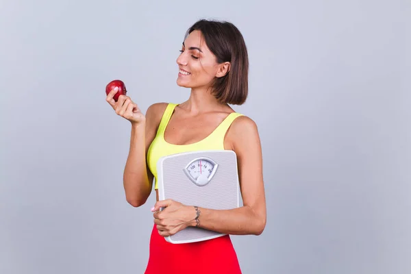 Spor kadını gri arka planda duruyor, fitness antrenmanı ve diyet sonuçlarından memnun, ölçeği tutuyor, üst ve tayt giyiyor, elma tutuyor.