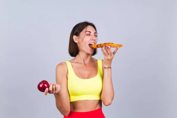 Spor kadını gri arka planda durur, fitness eğitimi ve diyet sonuçlarından tatmin olur, elmayı tutar ve kapalı gözlerle pizza yer, her lokmanın tadını çıkartır.