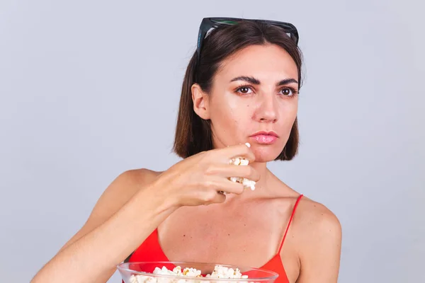 Beautiful woman on gray background in 3d cinema glasses looks aside focused on something having popcorn