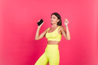 Pembe arka planda parlak spor kıyafetli güzel bir kadın. Kulaklarda dans eden kulaklıklar. Telefonu tut. 