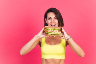 Pembe arka planda sarı parlak spor kıyafetli güzel bir kadın sağlıklı avokado sandviçi tutuyor.