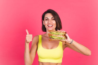 Pembe arka planda sarı parlak spor kıyafetli güzel bir kadın sağlıklı avokado sandviçi tutuyor.