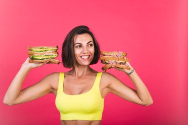 Pembe arka planda parlak spor kıyafetli güzel bir kadın. Sağlıklı avokado ve biftekli sandviç.