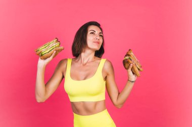 Pembe arka planda parlak spor kıyafetli güzel bir kadın. Sağlıklı avokado ve biftekli sandviç.
