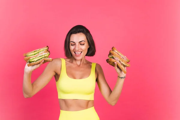 Beautiful fit woman in yellow bright fitting sportswear on pink background happy hold healthy avocado and roast beef sandwich