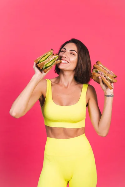 Beautiful fit woman in yellow bright fitting sportswear on pink background happy hold healthy avocado and roast beef sandwich