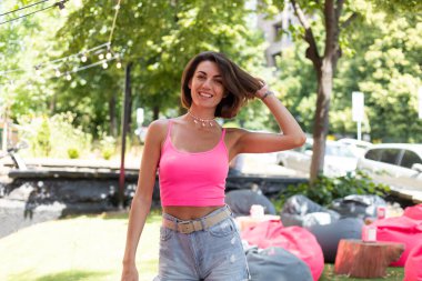 Parlak pembe bluzlu ve günlük kot şortlu güzel bir kadın yaz kafeteryasında boncuklu kolye takıyor.