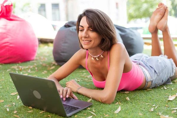 Beautiful woman in bright pink top and casual jean shorts wears beaded necklace in summer outdoor cafe on grass working on laptop