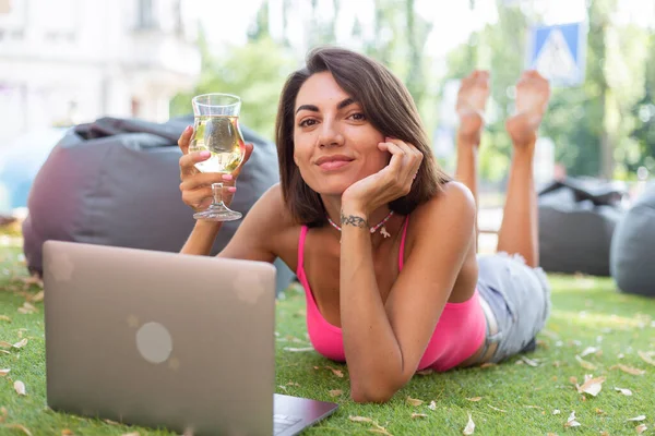 Beautiful woman in bright pink top and casual jean shorts wears beaded necklace in summer outdoor cafe on grass working on laptop