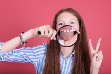 Pembe arka planda izole edilmiş 13 yaşındaki küçük kız çocuğu dişlerinin büyüteçle açıldığını ve dişlerinin diş tellerinin parantez içinde olduğunu gösteriyor.