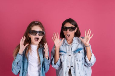 Terracotta arka planında kot ceketli, üç boyutlu sinema gözlüklü, korku filmi izleyen, korkmuş yüzlü modern anne ve kız.