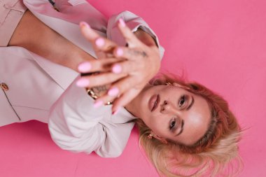 Zarif bej ceketli, altın takılı, güzel makyajlı, büyük dudaklı, pembe arka planda poz veren zarif Avrupalı bir kadın.