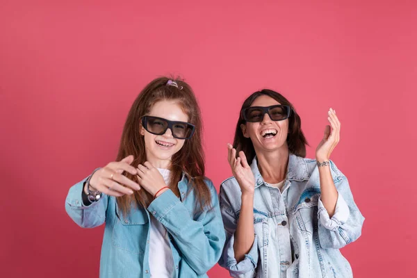 Modern mom and daughter in denim jackets on terracotta background in 3 d cinema glasses watching comedy movie together, smile and laugh