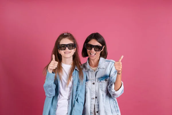 Modern mom and daughter in denim jackets on terracotta background in 3d cinema glasses positive smiling shows thumb up