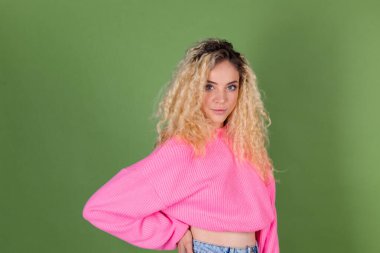 Uzun kıvırcık saçlı, yeşil arka planda pembe kazaklı, genç sarışın kadın kameraya gülümsüyor.