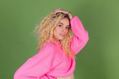 Uzun kıvırcık saçlı, yeşil arka planda pembe kazaklı, genç sarışın kadın kameraya gülümsüyor.
