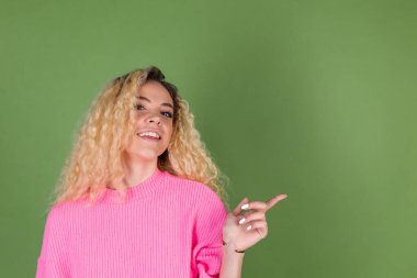 Uzun kıvırcık saçlı, yeşil arka planda pembe kazaklı, genç sarışın kadın. Sağdan sola gülümsüyor.