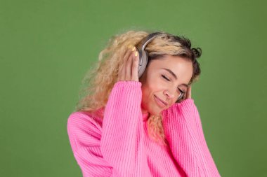 Yeşil arka planda pembe kazaklı, uzun kıvırcık saçlı genç sarışın kadın. Kablosuz kulaklıklarla müzik dinlemekten mutlu.