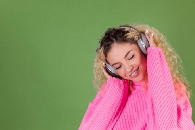 Yeşil arka planda pembe kazaklı, uzun kıvırcık saçlı genç sarışın kadın. Kablosuz kulaklıklarla müzik dinlemekten mutlu.