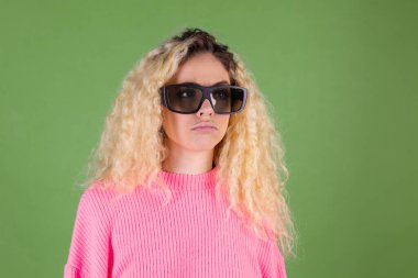 Uzun kıvırcık saçlı, pembe kazaklı, yeşil arka planlı, 3 boyutlu sinema gözlüklü, mutsuz, sarışın bir kadın.