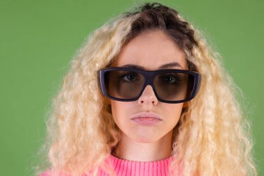 Uzun kıvırcık saçlı, pembe kazaklı, yeşil arka planlı, 3 boyutlu sinema gözlüklü, mutsuz, sarışın bir kadın.