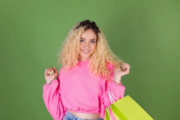 若いですブロンド女性とともに長いです巻き毛でピンクセーターで緑の背景にカラフルな紙ショッピングバッグ幸せな陽気興奮 — ストック写真