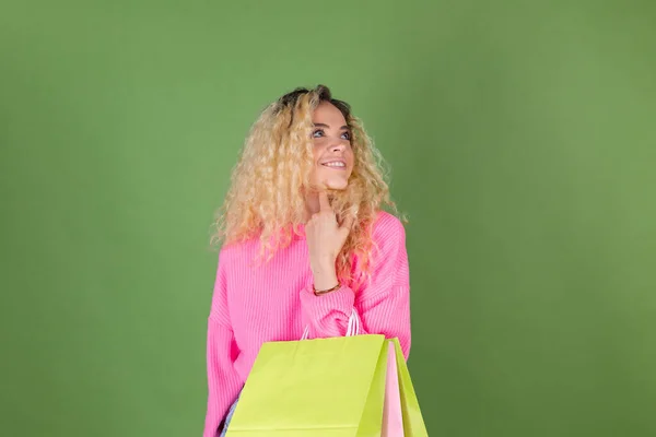 若いですブロンド女性とともに長いです巻き毛でピンクセーターで緑の背景にカラフルな紙ショッピングバッグ幸せな陽気な思慮深いです脇 — ストック写真