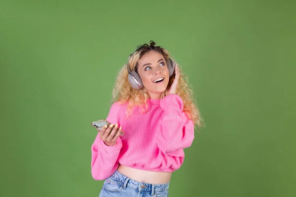若いですブロンド女性とともに長いです巻き毛でピンクセーターで緑の背景幸せな正楽しいですお気に入りの音楽で無線ヘッドフォン — ストック写真