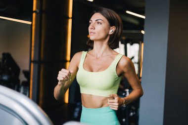 Fit caucasian beautiful woman in fitting sport wear at gym running jogging at treadmill with wireless headphones in ears