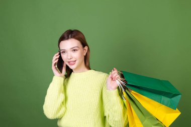Mükemmel makyajlı, kahverengi, yeşil arka planda koca dudaklı genç bir kadın alışveriş torbaları tutuyor. Cep telefonunda heyecanlı konuşmalar yapıyor.